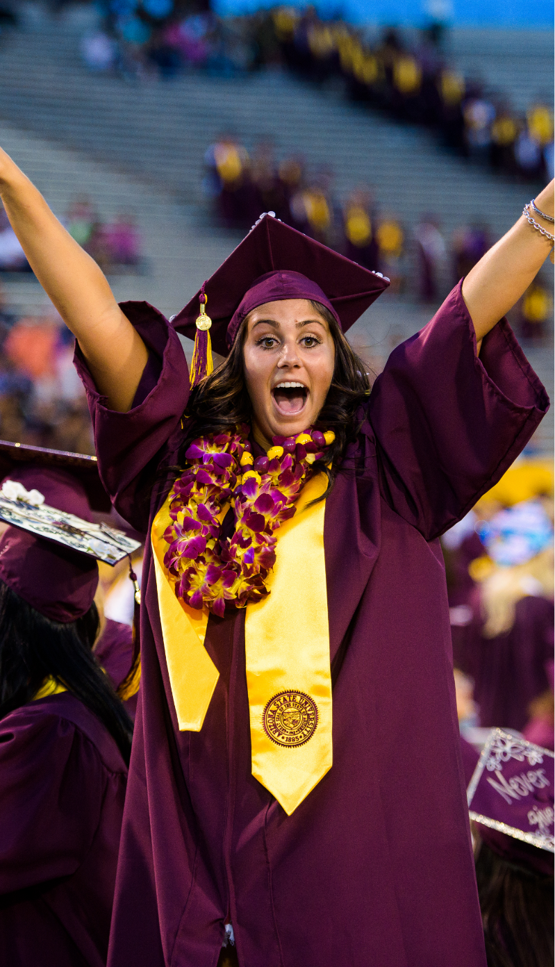 At graduation, an ASU student celebrates in her cap and gown with her arms up high.